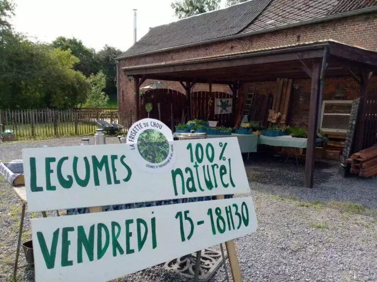 Un marché rustique en plein air avec une enseigne annonçant des produits et légumes « 100% naturels » en vente sous un abri en bois, dans un environnement champêtre, mettant en avant l'offre des maraîchers locaux.