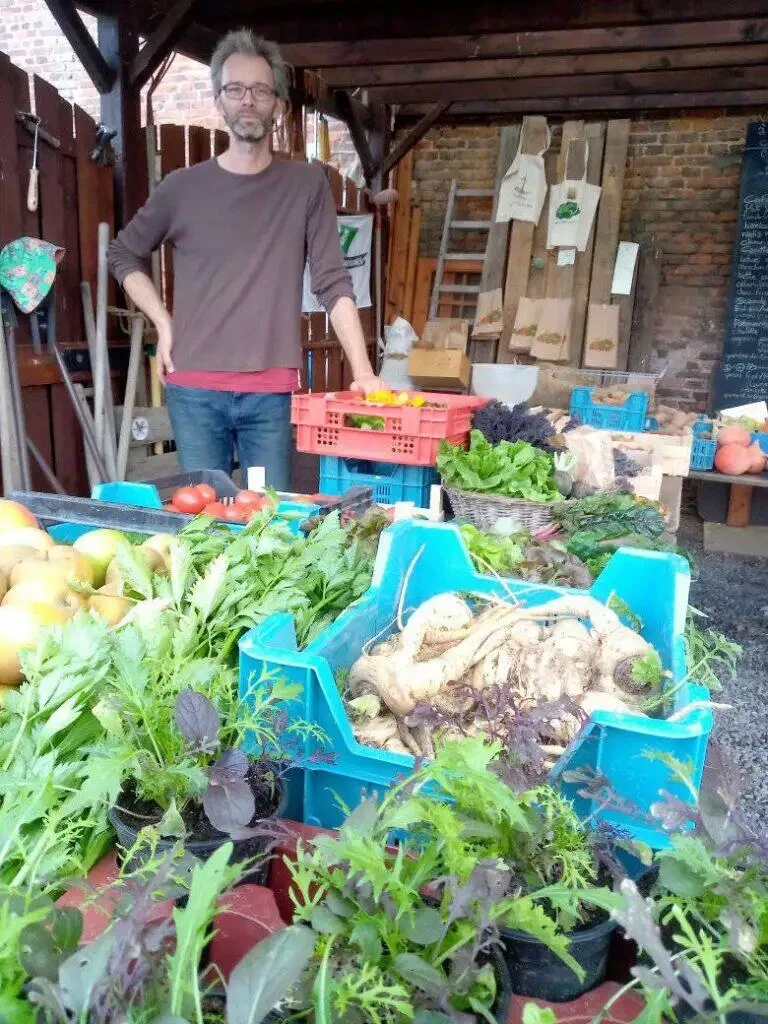 Homme debout derrière une table remplie de légumes frais et de plantes sur un étal de marché maraîcher.