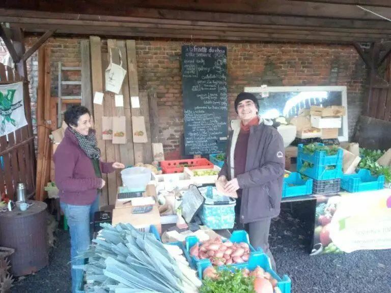 Deux personnes devant un stand de maraîcher rustique en plein air rempli de légumes frais locaux et d’un menu au tableau.