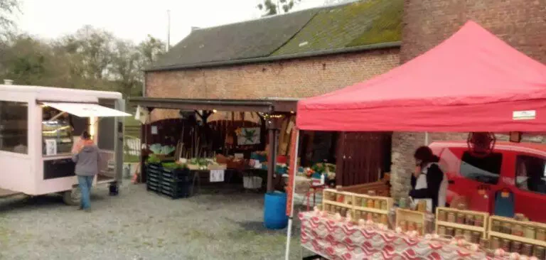 Marché en plein air avec vendeurs de produits et marchandises sous tentes et en arrière-plan un bâtiment de style grange à Blarégnies.