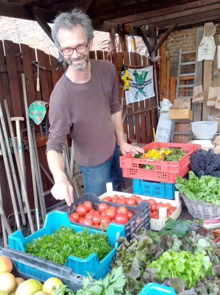 Un homme aux lunettes et aux cheveux grisonnants se tient derrière des caisses de légumes frais de son abonnement maraîcher, souriant à la caméra dans un décor extérieur rustique.