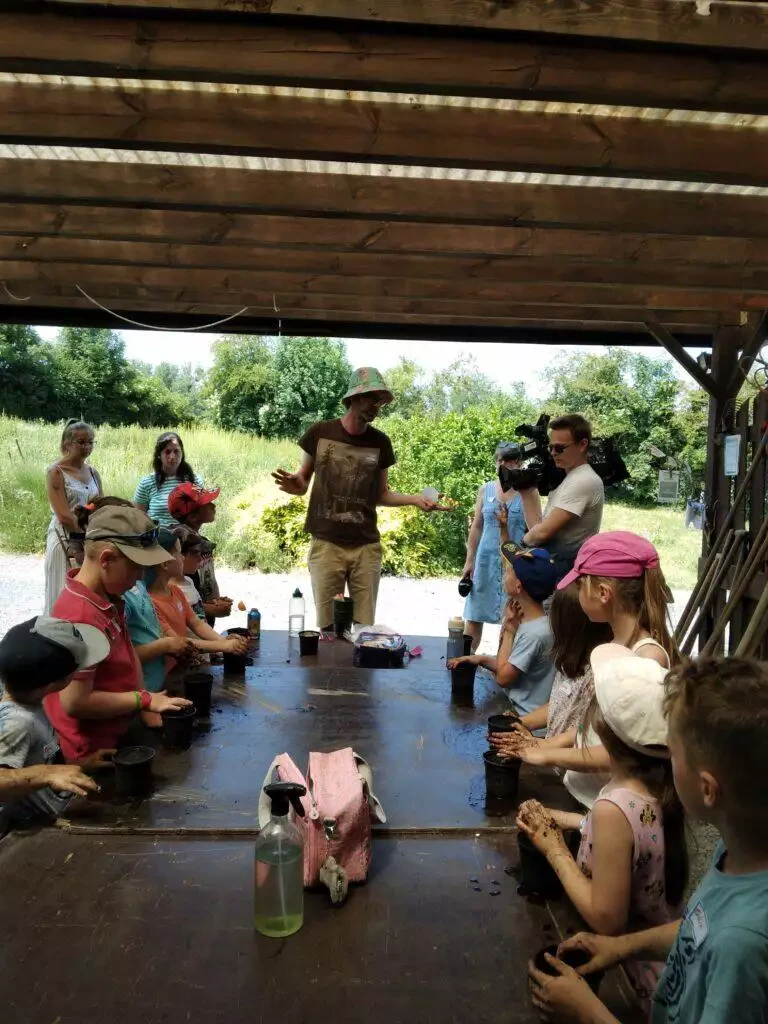 Un groupe de personnes, comprenant des enfants et des adultes, participant à un atelier animé par un homme local portant un chapeau, sous un espace extérieur abrité.