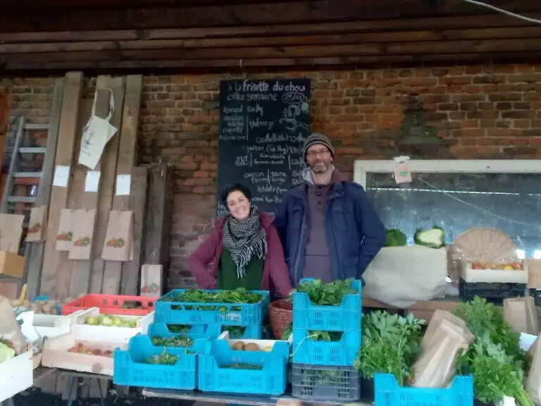 Deux personnes debout derrière un étal de marché rempli de légumes frais, avec un menu au tableau en arrière-plan.