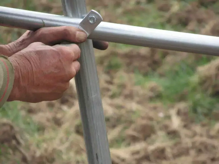 Gros plan d'une main patinée saisissant une structure de tuyaux métalliques à l'extérieur, avec un fond de végétation locale floue.