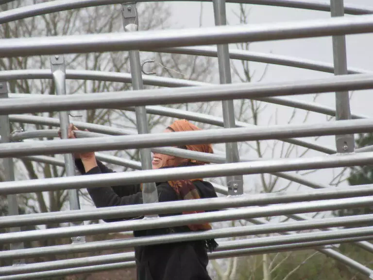 Une personne ajustant des échafaudages métalliques à l’extérieur, souriante pendant qu’elle travaille, sur un fond flou de légumes verdoyants.