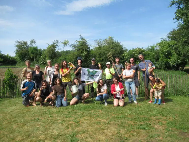 Un groupe de locaux heureux d'âges divers posant à l'extérieur dans une zone herbeuse, tenant un drapeau, avec des arbres et un ciel dégagé en arrière-plan.