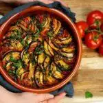Une cocotte remplie de ratatouille colorée en couches à côté de tomates fraîches locales sur une surface en bois, partiellement tenue à la main avec un chiffon.