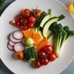 Une assiette blanche avec une variété de légumes crus locaux, notamment des tomates, du brocoli, des carottes, des oignons et des courgettes sur fond gris.